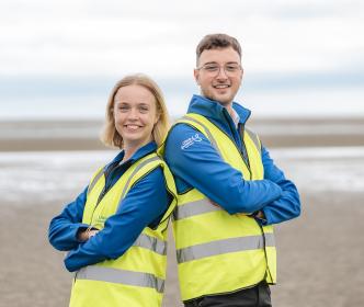 Two graduates folding their arms wearing a Uisce Éireann jacket with a high visible jacket on a beach 